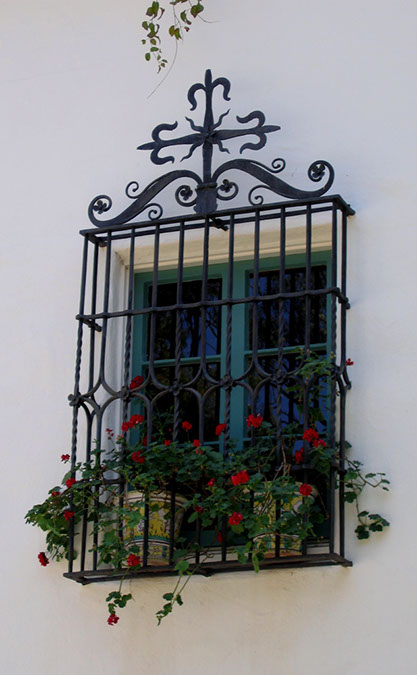 An ornate wrought iron window grille with scrolling and cross with pots and red geraniums at Casa del Herrero estate in Montecito, California
